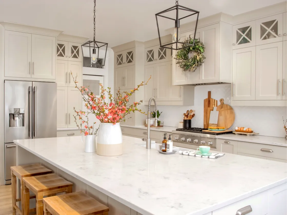 An elegant farmhouse-style kitchen with soft beige cabinets and a large marble island complemented by decorative greenery