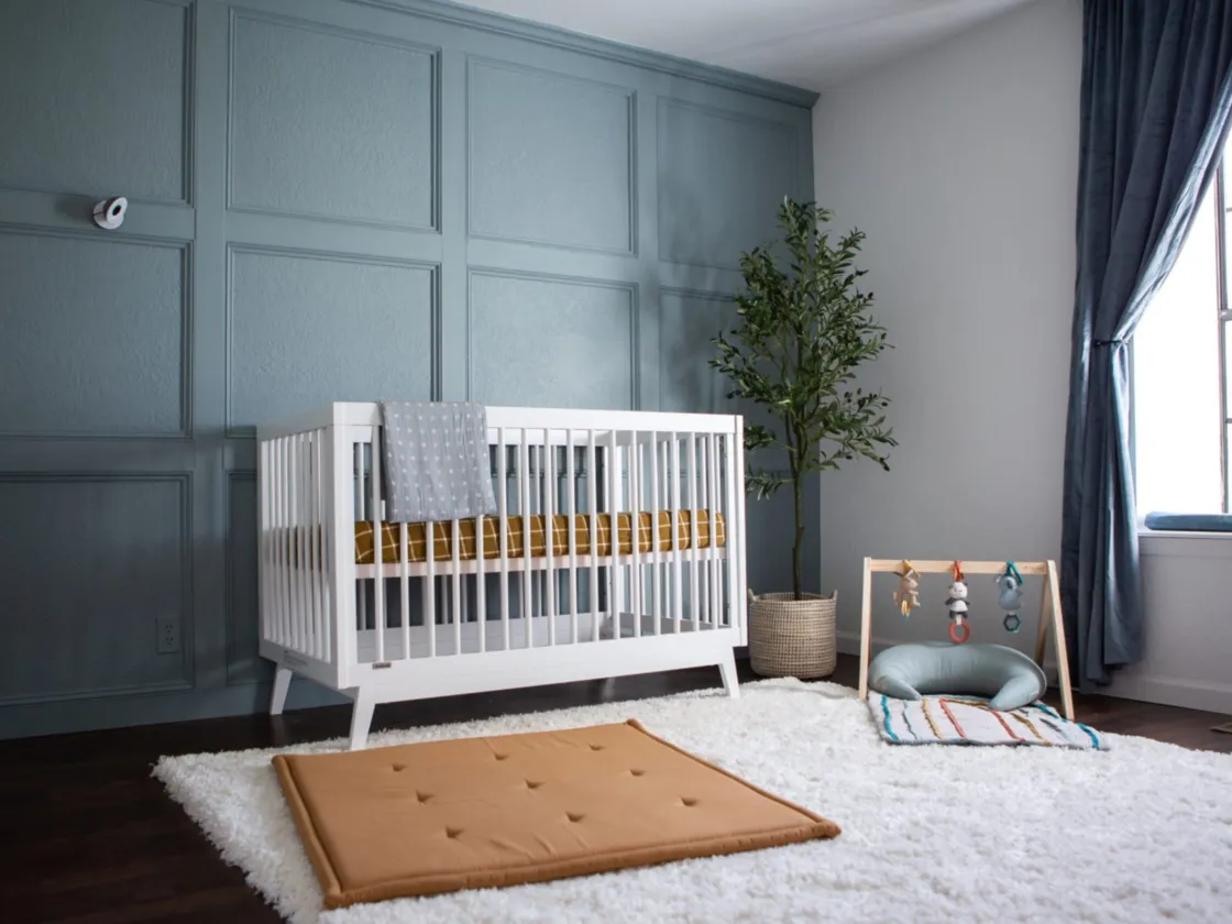 A modern gender-neutral nursery with a muted blue-gray accent wall, white crib, cozy rug and mustard accents
