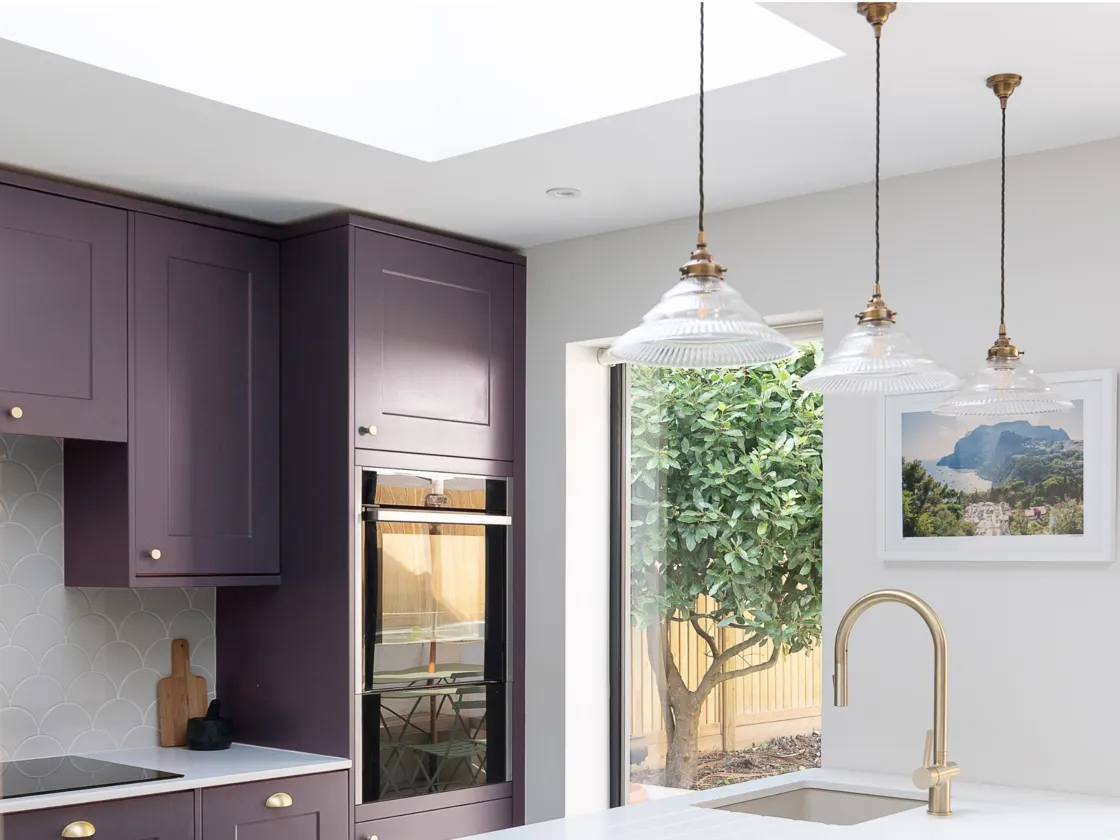 A bright kitchen space with deep purple cabinets, crisp white walls and gold accents that overlooks the lush garden outside